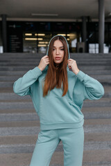 Fototapeta premium Cute girl model in a sporty comfortable casual turqouise outfit with a hoodie and sweatpants posing on the street
