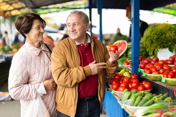 Positive senior couple in casual wear picking tomatoes during shopping at crowded bazaar