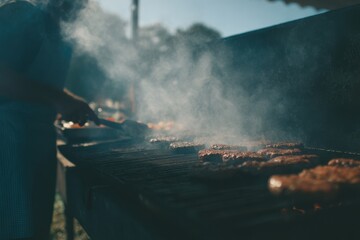 Flipping burgers on a grill amidst smoke on a sunny day at an outdoor cooking event