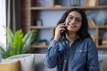 A happy young woman wearing glasses smiles while talking on her mobile phone at home.