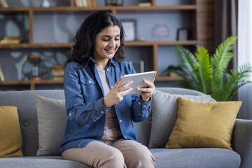 A cheerful young woman is engrossed in her tablet while relaxing on a comfy sofa at home.