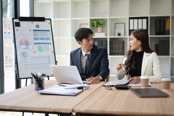 Businessman and businesswoman discussing strategy and financial reports at a desk with laptop and charts. Professional office teamwork in a bright, organized workspace