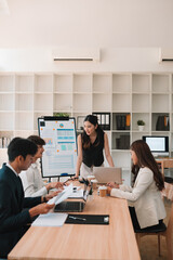 A team of young professionals engages in a strategy meeting in a bright office. A woman stands leading the discussion while others focus on charts, documents, and laptops