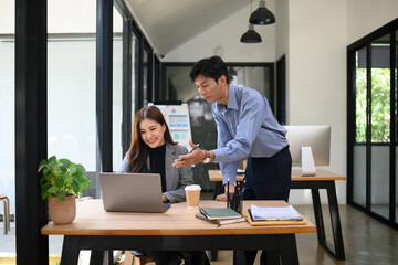 Smiling businesswoman working on a laptop while a male colleague points at the screen, discussing ideas together in a bright, modern office, Concept of teamwork and support