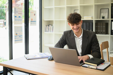 Young Caucasian businessman smiling while working on a laptop in a bright modern office. Paperwork, notebook, and coffee cup on desk, bookshelves in background