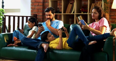 Indian family using smartphones individually while sitting together on living room couch, showing screen addiction in modern lifestyle, no interaction or bonding, lack of quality family time concept - Powered by Adobe