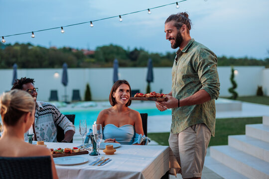 Man serving appetizers to friends dining by the pool during a summer evening party - Powered by Adobe