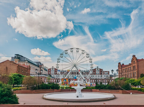 Ferris wheel and a non-operational fountain in a town square in Żyrard&oacute;w, Poland, surrounded by brick buildings under a partly cloudy sky.