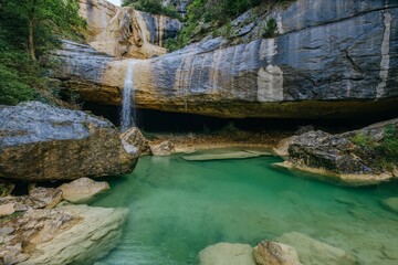 Enchanting cave waterfall flowing into pristine jade pool. Natural limestone grotto with emerald waters forming secret swimming hole beneath massive rock overhang. Pozas de San Martin, Huesca