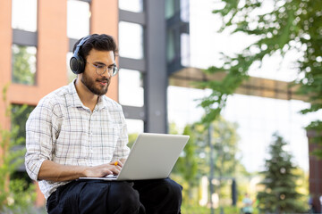 Fototapeta na wymiar Young student wearing headphones and using a laptop, studying outdoors on campus.