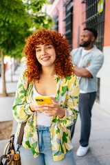 Smiling young woman with curly red hair using mobile phone on a city street