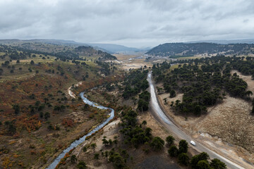 Winding road and stream cut through hills dotted with Araucaria trees under a cloudy sky. Mountains...