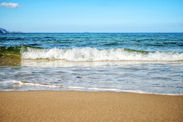 Calm ocean wave approaches smooth sandy shore beneath clear blue sky. Tranquil seascape highlights cresting water, shimmering reflections, and distant horizon in bright sunlight.