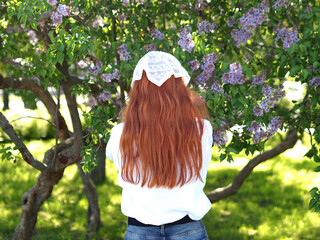 Portrait of a beautiful red-haired young woman in a headscarf in lilac blossom blossoms. Back view