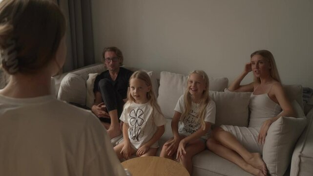 A family is having a blast playing charades at home. One daughter energetically acts out clues while her parents and sisters guess with great excitement, one girl even jumping with joy.