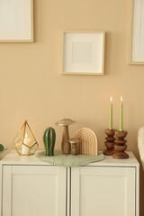 Stylish candlesticks with burning candles and other decor on cabinet near beige wall indoors