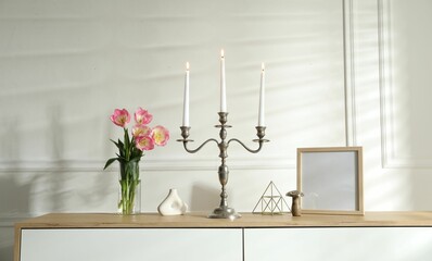 Elegant candlestick with burning candles and decor on chest of drawers near white wall