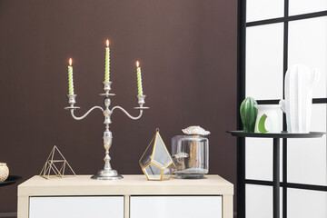 Elegant candlestick with burning candles and decor on cabinet near brown wall
