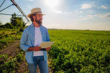Fototapeta premium Confident farmer holding a digital tablet while admiring crops in a cultivated field with an irrigation system, highlighting modern technology for sustainable agriculture