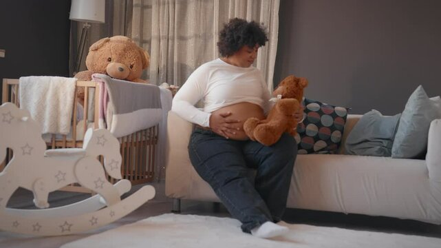African american woman sitting alone in cozy apartment, stroking pregnant belly. Lady looking at teddy bear, waiting for her first baby, eighth month of pregnancy, firstborn in family, motherhood