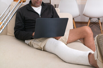 Man with broken leg using laptop on sofa with crutches nearby