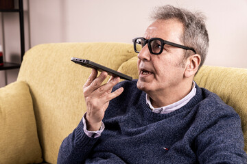 Senior man using smartphone voice assistant while sitting on couch at home