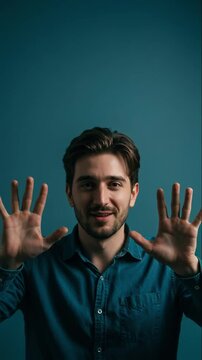 Cheerful young man in a blue shirt smiles widely showing ten fingers with open palms against a blue background, indicating a count of ten or expressing playful excitement for concepts of quantity