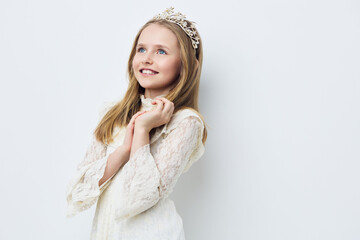 Sweet young girl with a tiara in a white dress smiling happily against a soft white background,...