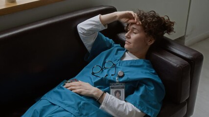 Young tired female doctor in medical scrubs putting off eyeglasses while relaxing on leather couch in break room after hard working day and trying to get asleep