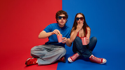 Young man and woman, couple sitting on the floor wearing 3D glasses and eating popcorn, with surprised expressions, against vivid red and blue background. Concept of movies, reaction, emotions