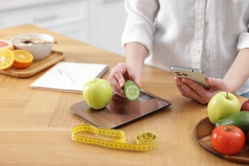 Woman with smartphone weighting products on kitchen scale at wooden table, closeup
