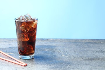 Refreshing cola with ice cubes in glass and straws on grey textured table against light blue...