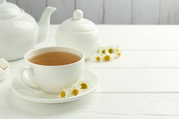 Delicious chamomile tea in cup, fresh flowers and teapot on white wooden table, closeup. Space for text