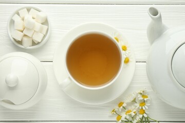 Delicious chamomile tea in cup, fresh flowers, sugar cubes and teapot on white wooden table, flat lay