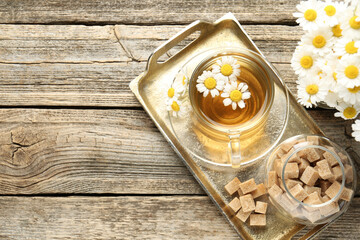 Delicious chamomile tea in glass cup, fresh flowers and brown sugar on wooden table, flat lay. Space for text
