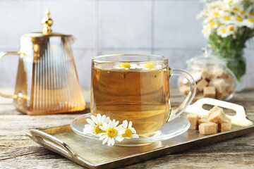 Delicious chamomile tea in glass cup, fresh flowers, brown sugar and teapot on wooden table, closeup