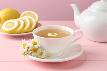 Delicious chamomile tea in cup, fresh flowers and lemon on pink wooden table, closeup