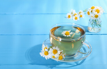 Delicious chamomile tea in glass cup and fresh flowers on light blue wooden table, closeup. Space for text