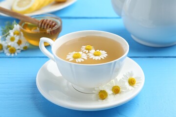 Delicious chamomile tea in cup, fresh flowers and honey on light blue wooden table, closeup