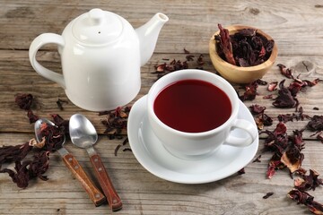 Delicious hibiscus tea in cup, teapot, dry roselle sepals and spoons on wooden table