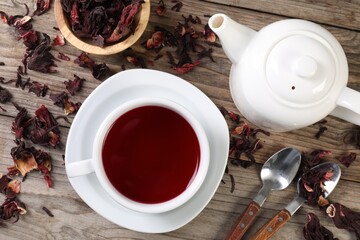 Delicious hibiscus tea in cup, teapot, dry roselle sepals and spoons on wooden table, flat lay