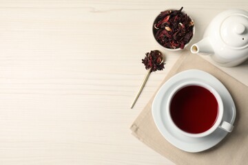 Delicious hibiscus tea in cup, teapot, dry roselle sepals and spoon on light wooden table, flat...