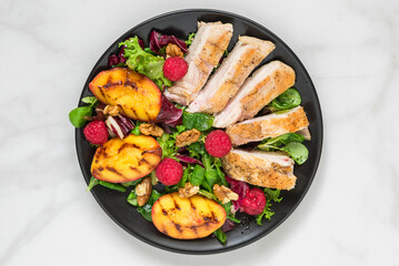 Chicken breast fillet salad with grilled peaches, raspberries, walnuts and mixed greens on black plate on white marble background. Top view. Healthy diet food. Summer lunch
