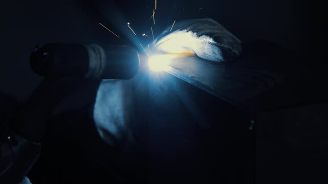 Welder performing TIG welding with bright arc light, Close-up of gloved hands joining metal in dark workshop environment