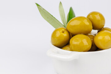 Closeup of green olives in white bowl and olive branch on white surface