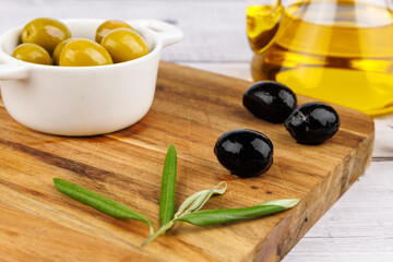 Glass olive oil bottle with green and black olives and olive leaves on dark board and white...