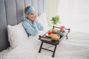 Young woman in pajamas enjoys breakfast in bed with a surprised expression during a relaxing...
