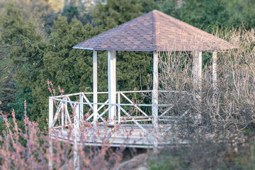 Wooden gazebo in park setting, late afternoonearly evening, shingled roof, vertical posts, railing,...