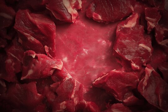 A close up of a pile of meat with a red background. The meat is cut into small pieces and he is raw. The image has a raw and primal feel to it, evoking a sense of hunger and primal instincts