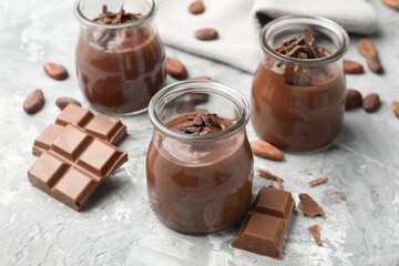 Tasty pudding in glass jars, pieces of chocolate and cocoa beans on light grey table, closeup